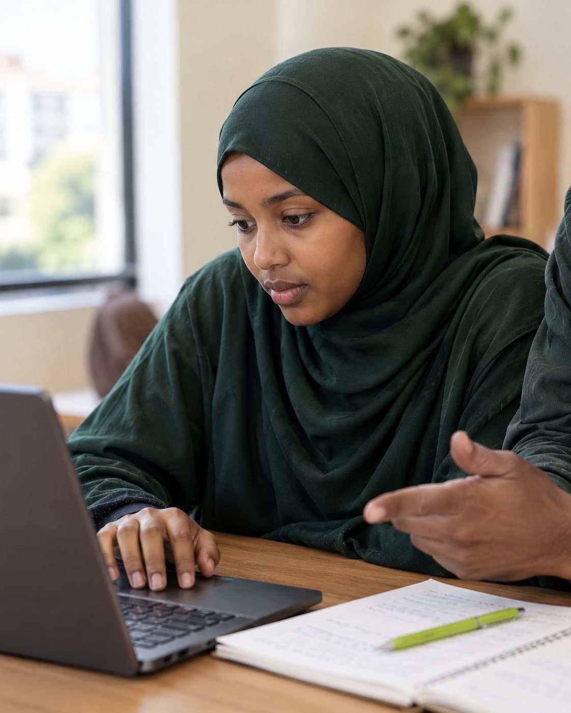 A young woman works on a laptop while a mentor guides her